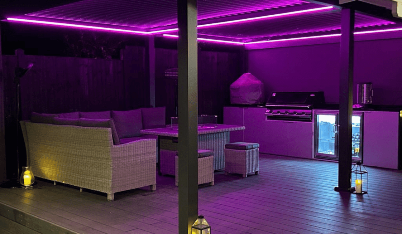 Outdoor entertaining area and kitchen under purple LED light from the pergola. Featuring an outdoor seat and table and straight run of kitchen