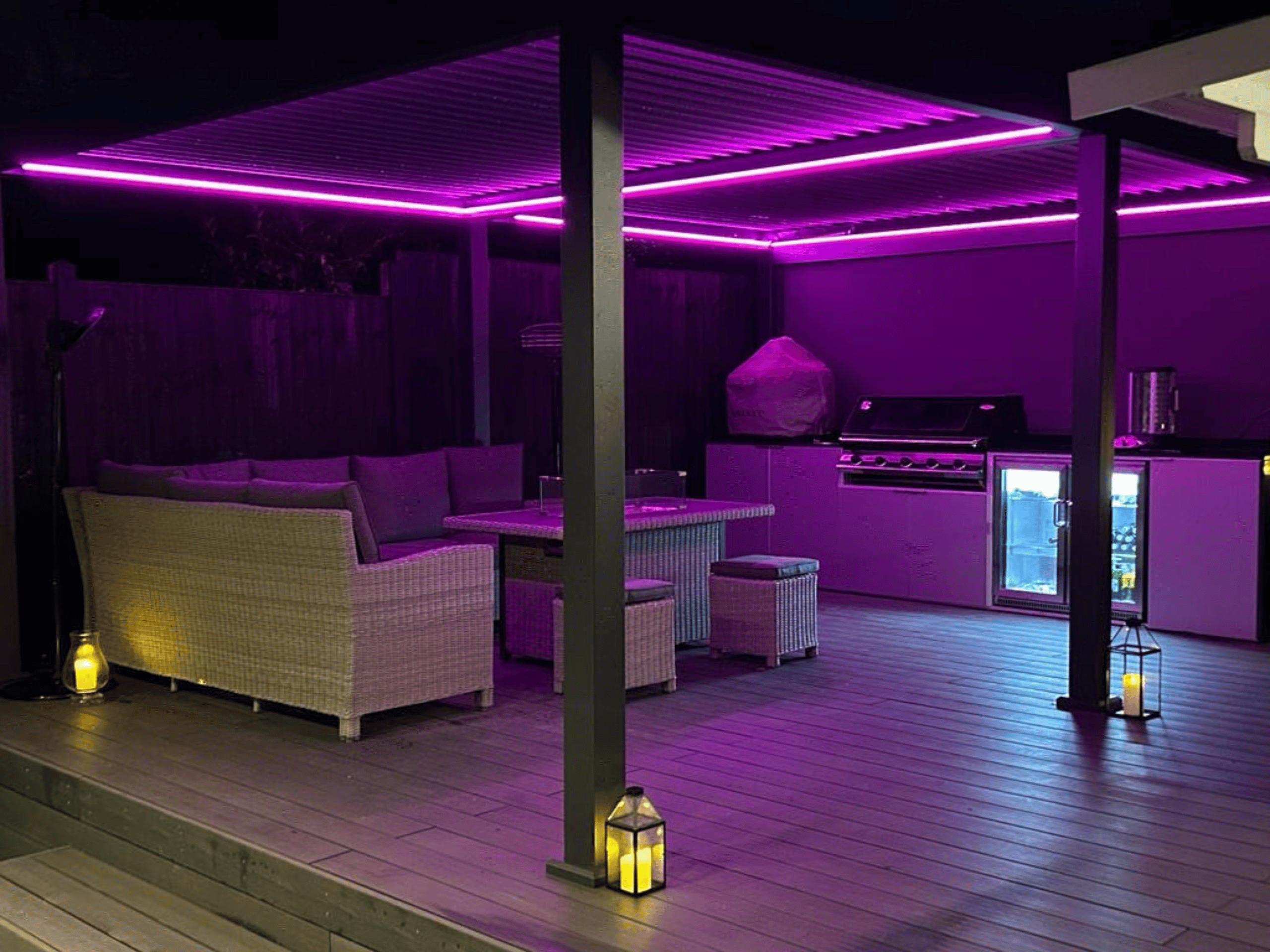 Outdoor entertaining area and kitchen under purple LED light from the pergola. Featuring an outdoor seat and table and straight run of kitchen