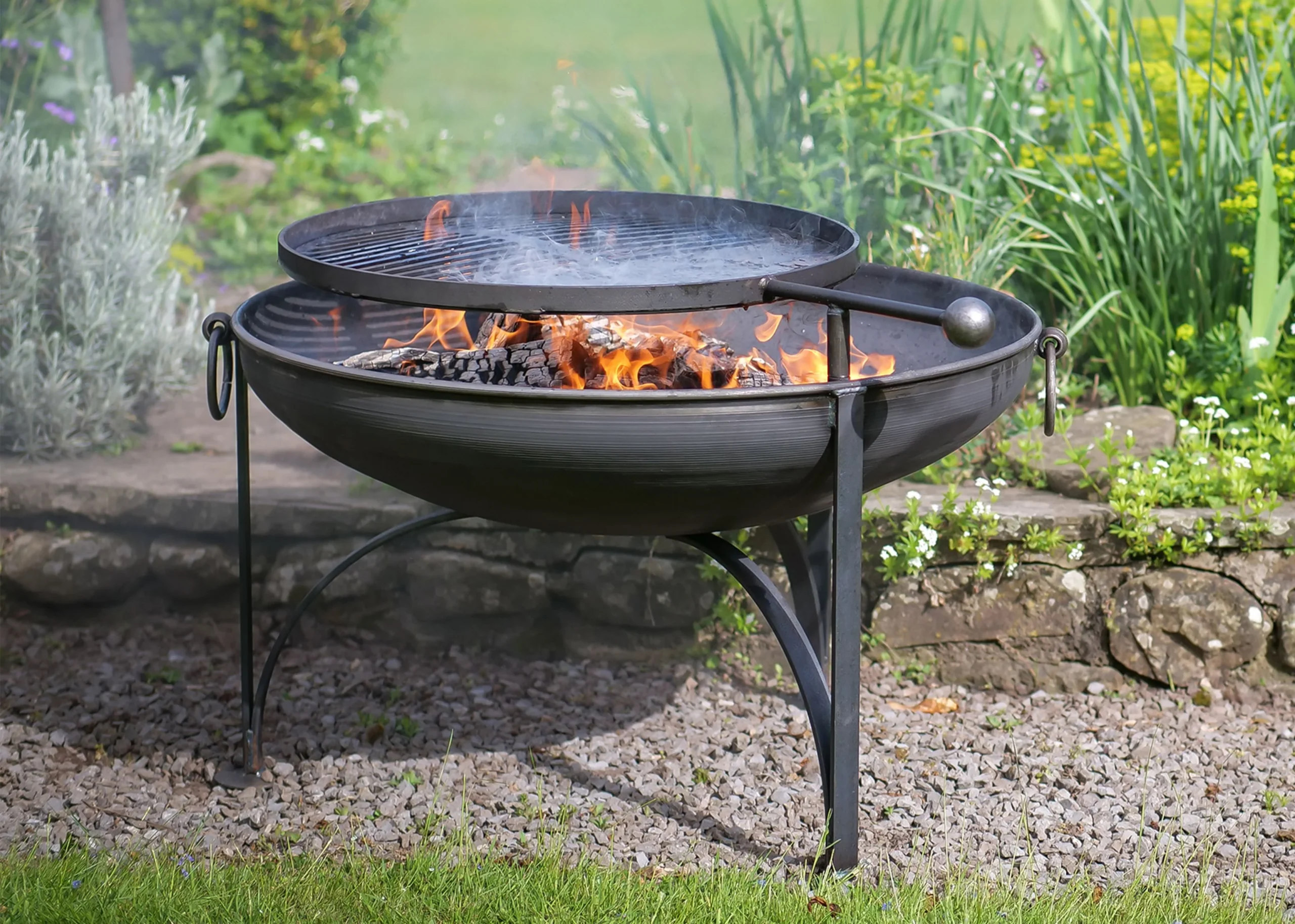 Black metal firepit with swing arm BBQ attachement. The fire pit is in a green garden environment with lit coals.