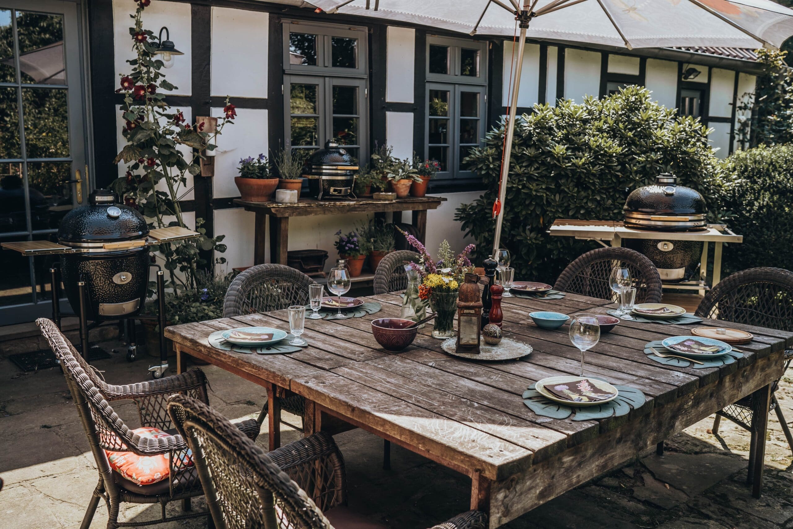 Outdoor dining area with a rustic wooden table set for a meal, surrounded by various sized Monolith ceramic barbecue grills and potted plants.