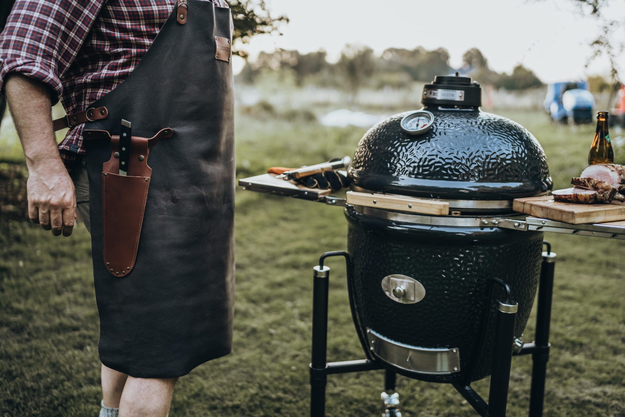 Man in a leather apron standing next to the monolith black kamado BBQ which is closed with side table wings out.