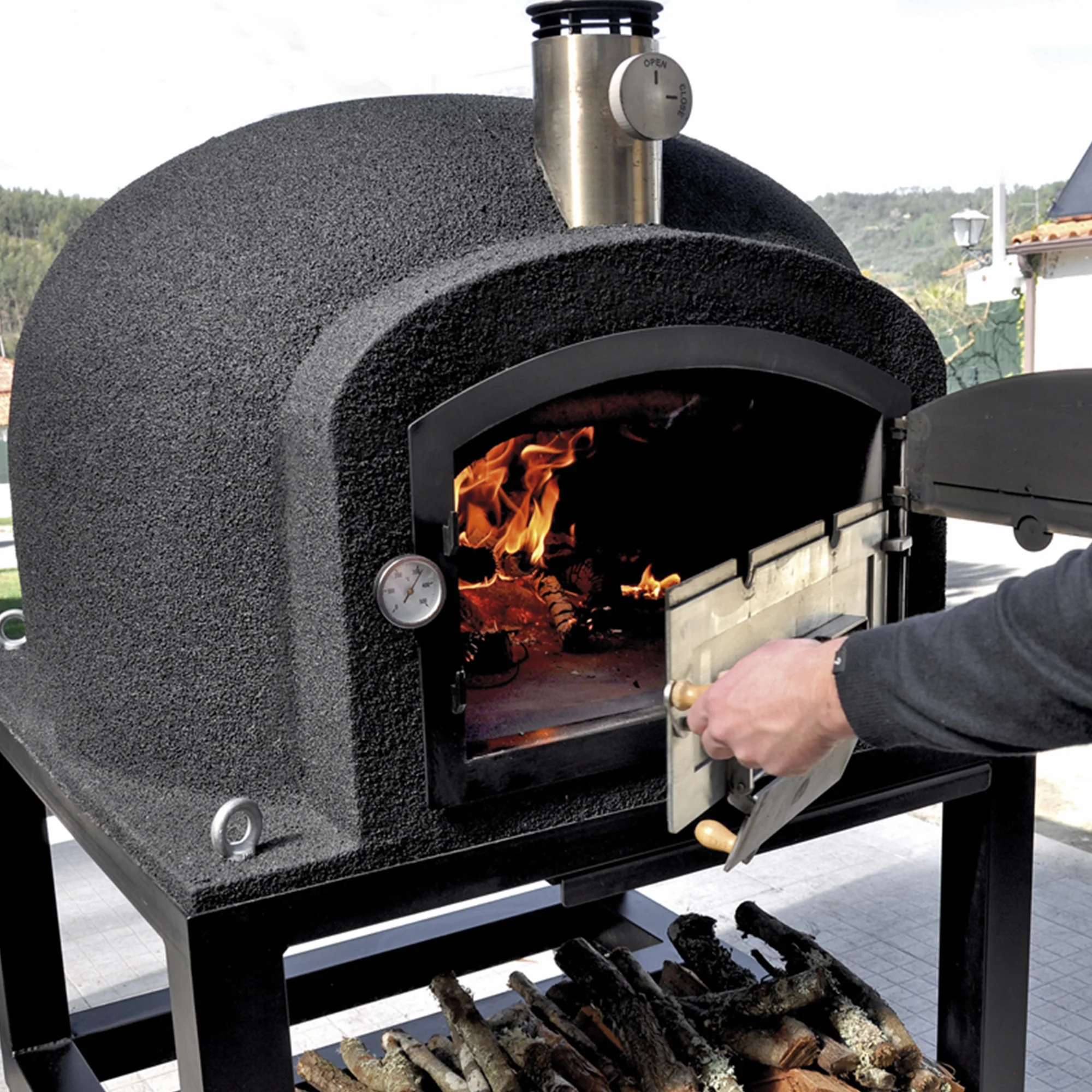 Large Tuozi wood fired oven being opened by a person as a fire is roaring inside. The oven stand on its moveable metal stand,