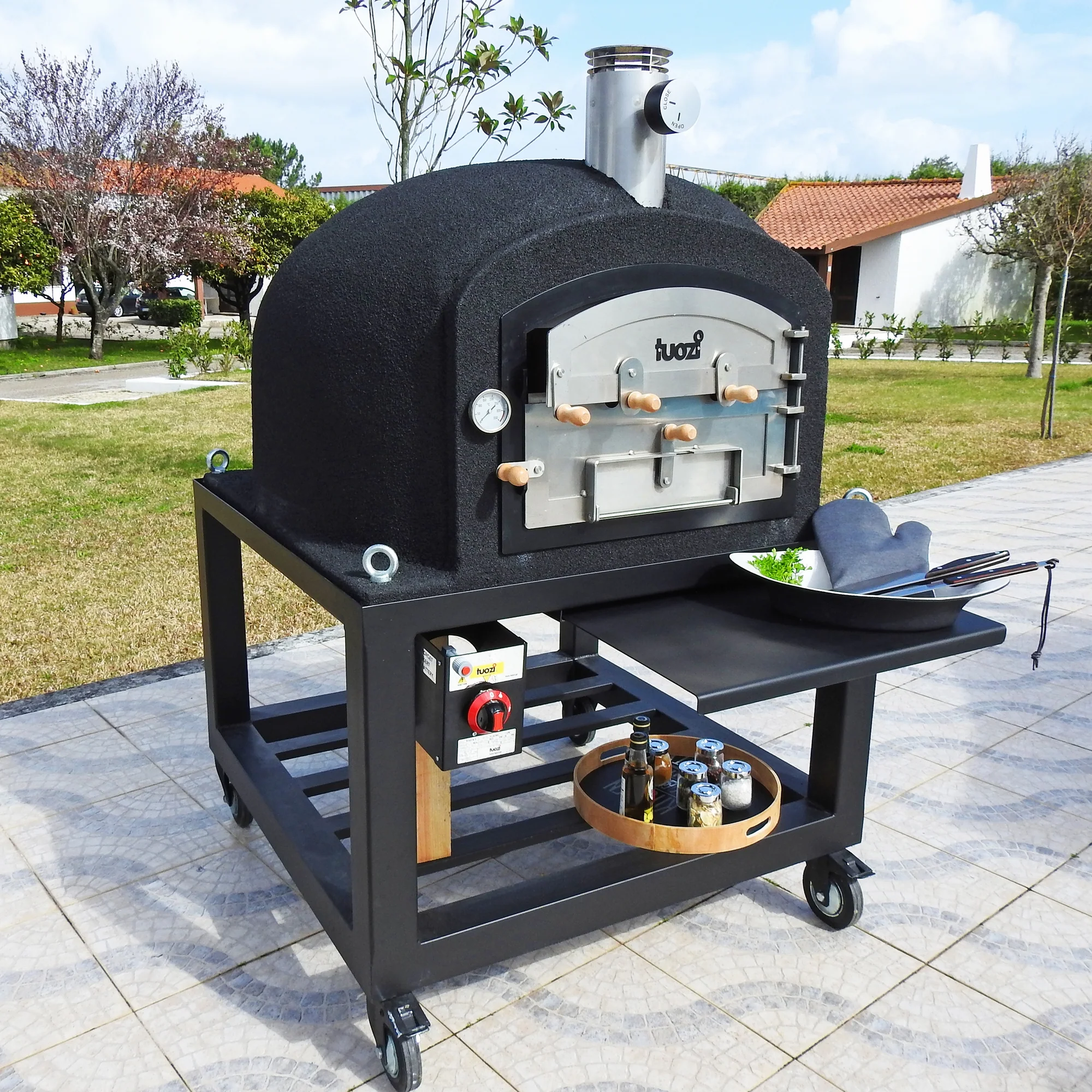 Black Tuozi Wood Fired Pizza Oven on its movable black trolley in a garden using the pull out front tray to hold food ready for cooking.