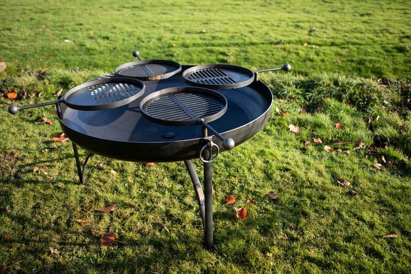 A brand new empty black metal fire pit with 4 swin arm grills in the setting of a field.