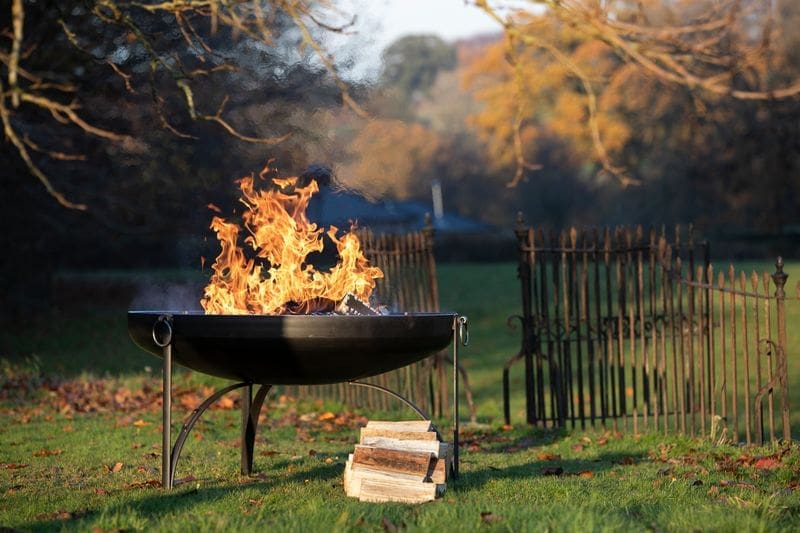 Black firepit with a fire burning well. With a pile of logs next to the fire pit the setting is the countryside in Autumn