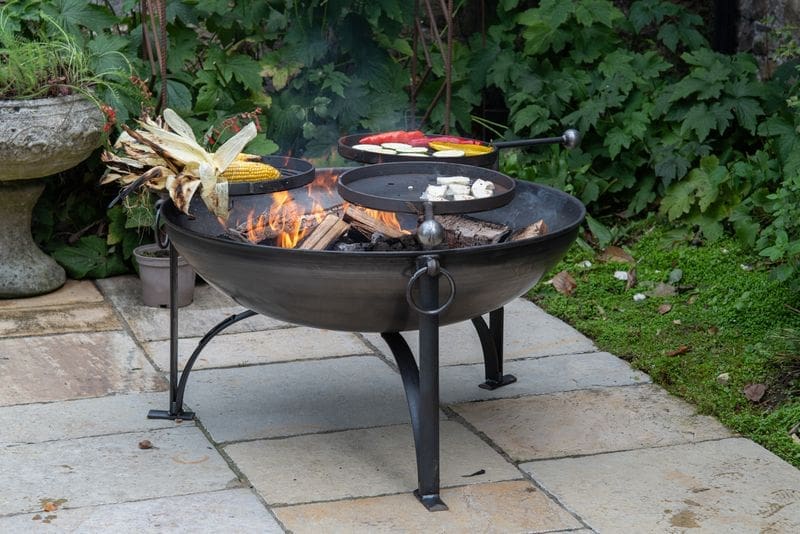 Classic 90cm black metal fire pit with 3 swing arms cooking food on a garden patio.