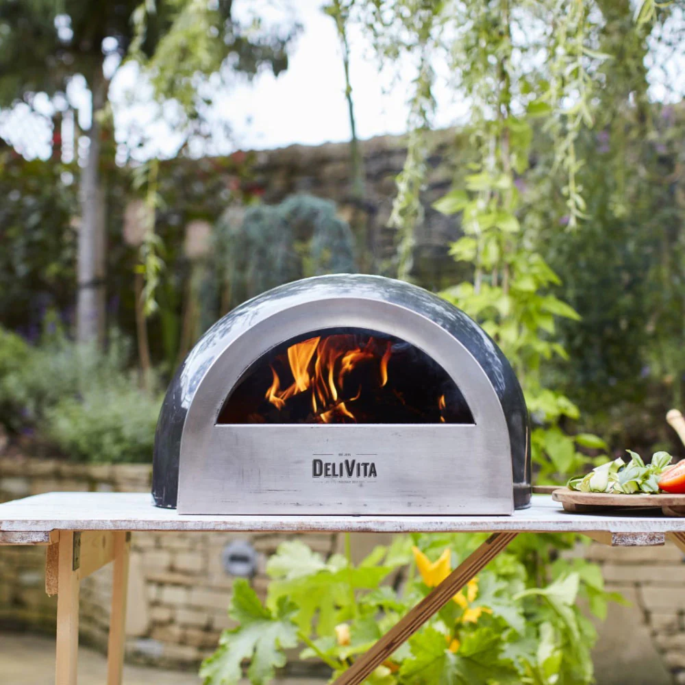 Grey Pizza oven sat on a wooden table top in the garden with a plate of food to one side and fired burning inside