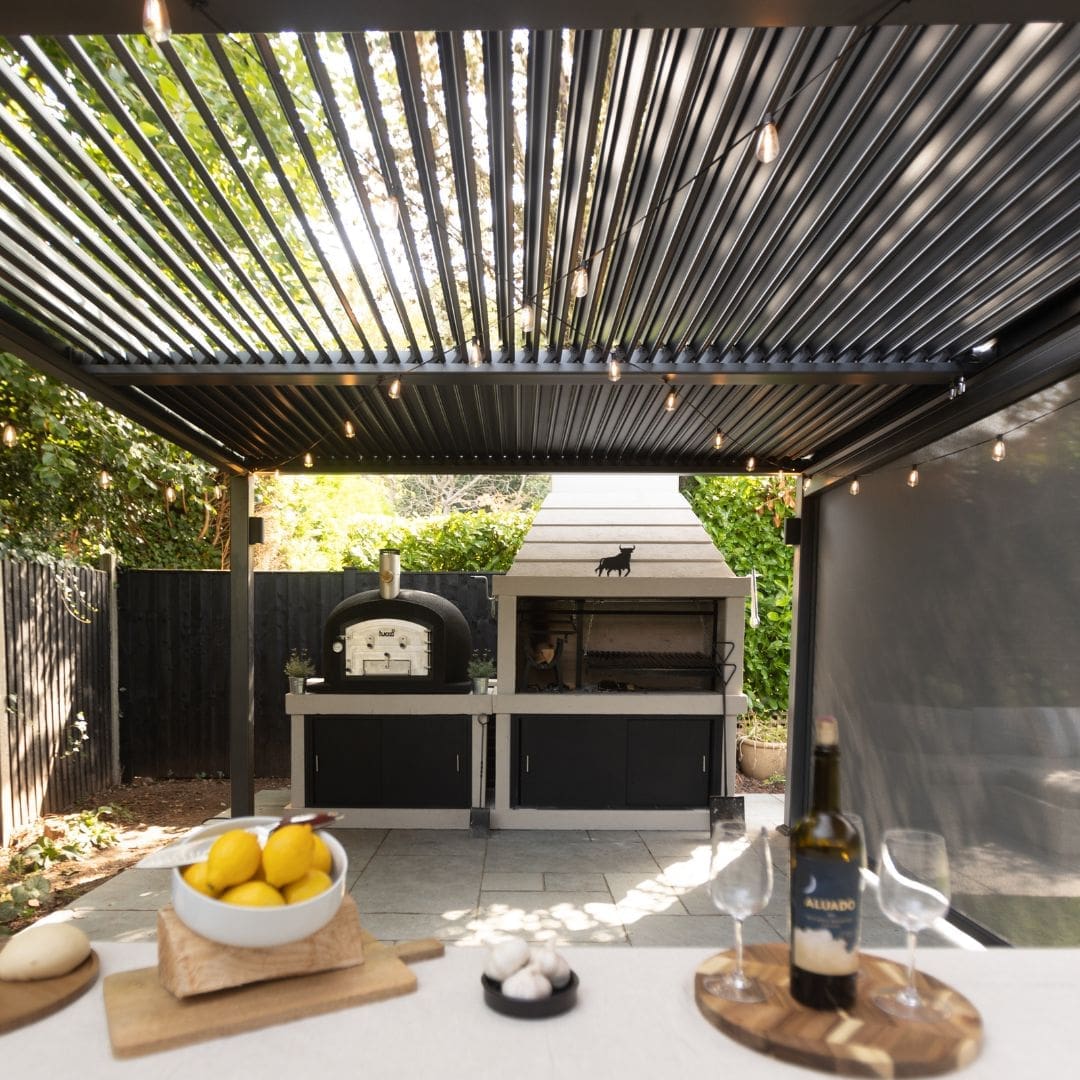 Outdoor kitchen with an Argentinian precast concrete barbecue grill and a black wood-fired oven under an anthrocite metal louvred pergola.