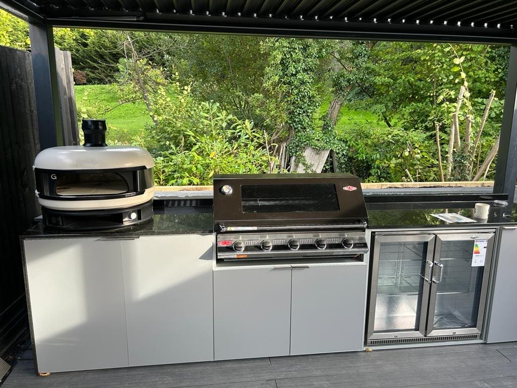 Gozney dome on an outdoor kitchen worktop with grey doors and a beefeater BBQ and fridge. The kitchen overlooks green countryside